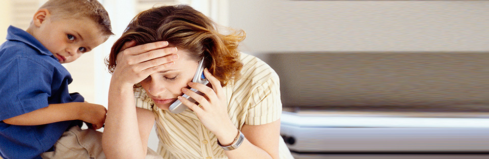 A  stressed woman talks on a phone while holding her head, with a young child sitting on the counter beside her.