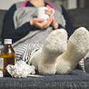 Woman sitting on couch with feet propped up on ottoman. A bottle of cold medicine is sat beside her feet.