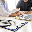 Doctor and patient seated at table in front of a laptop, with stethoscope and clipboard placed off to the side.